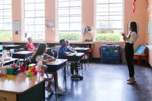 Students sit at desks in a class listening to a teacher