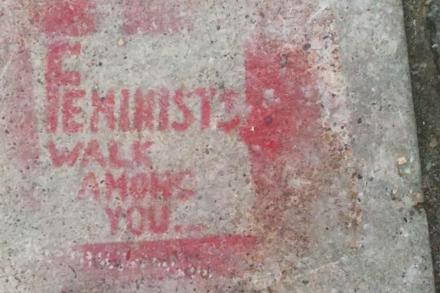 A light grey concrete sidewalk is spray painted with the phrase "Feminists Walk Among You."