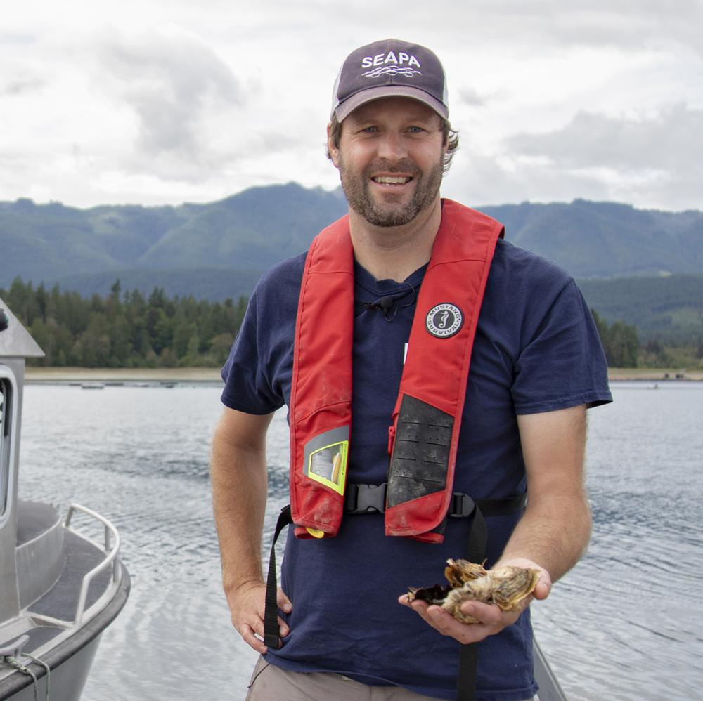 Dr. Timothy Green, VIU’s Canada Research Chair in Shellfish Health and Genomics stands on a dock next to a boat holding oysters in his hand.