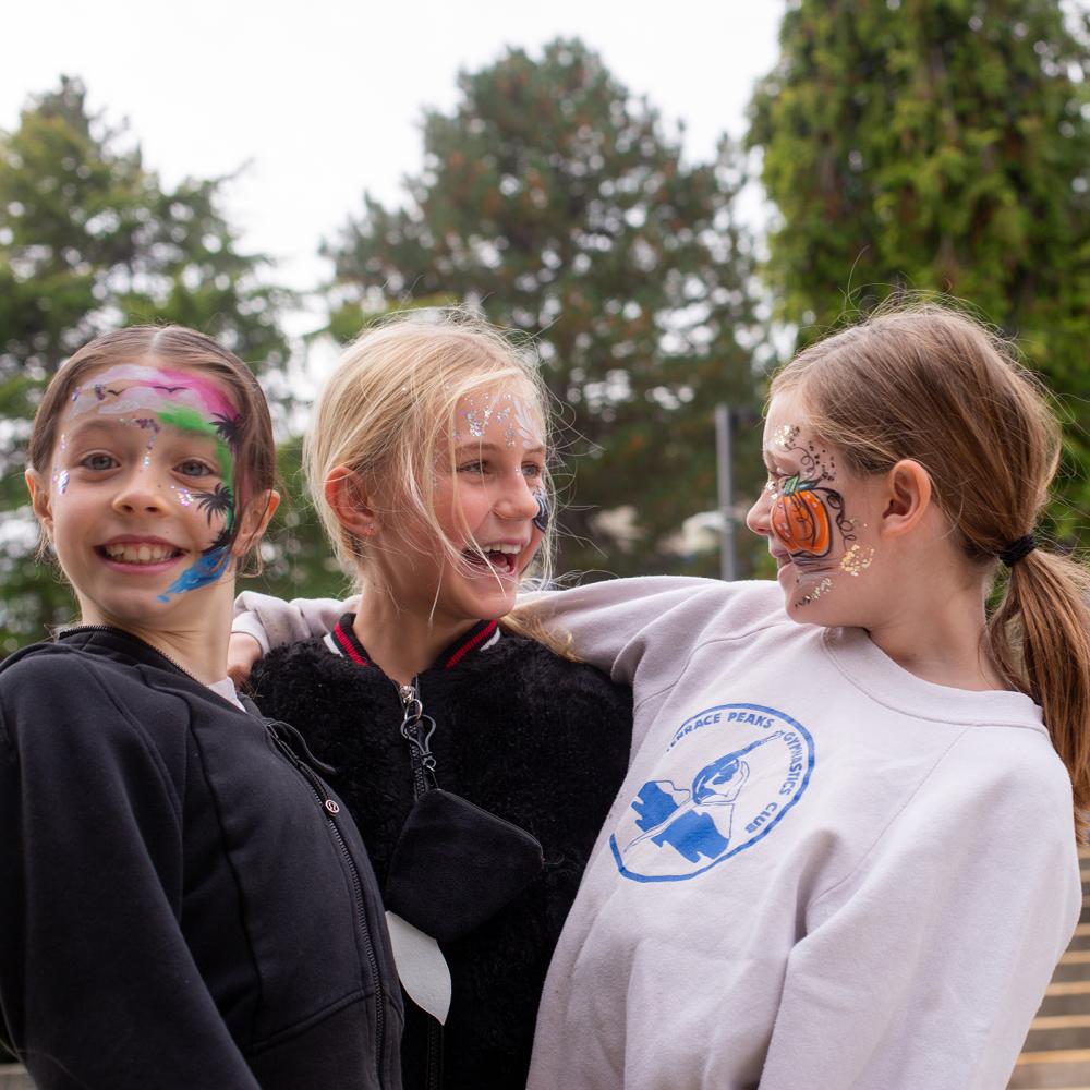 Three young girls wearing face paint hug, with a crowd on the stairs behind them