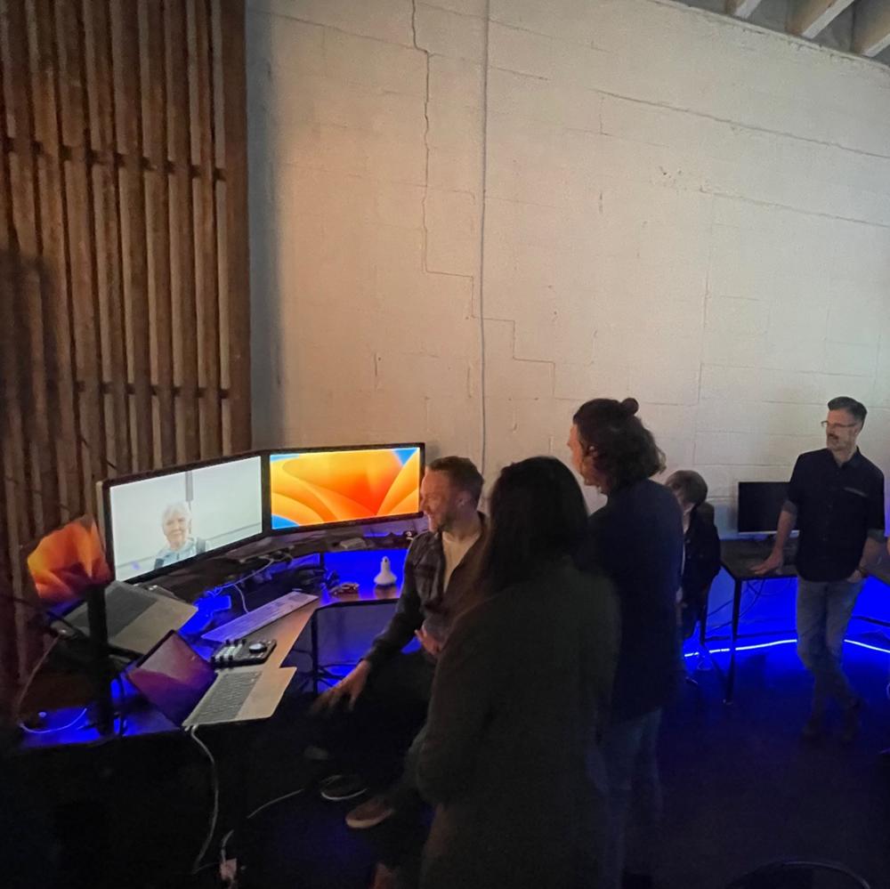 Students gather around three screens on a desk with a projection of an image on the wall behind them.